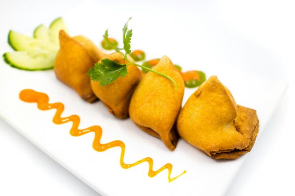 Appetizing Indian samosas served with cucumber slices and sauce on a white plate.